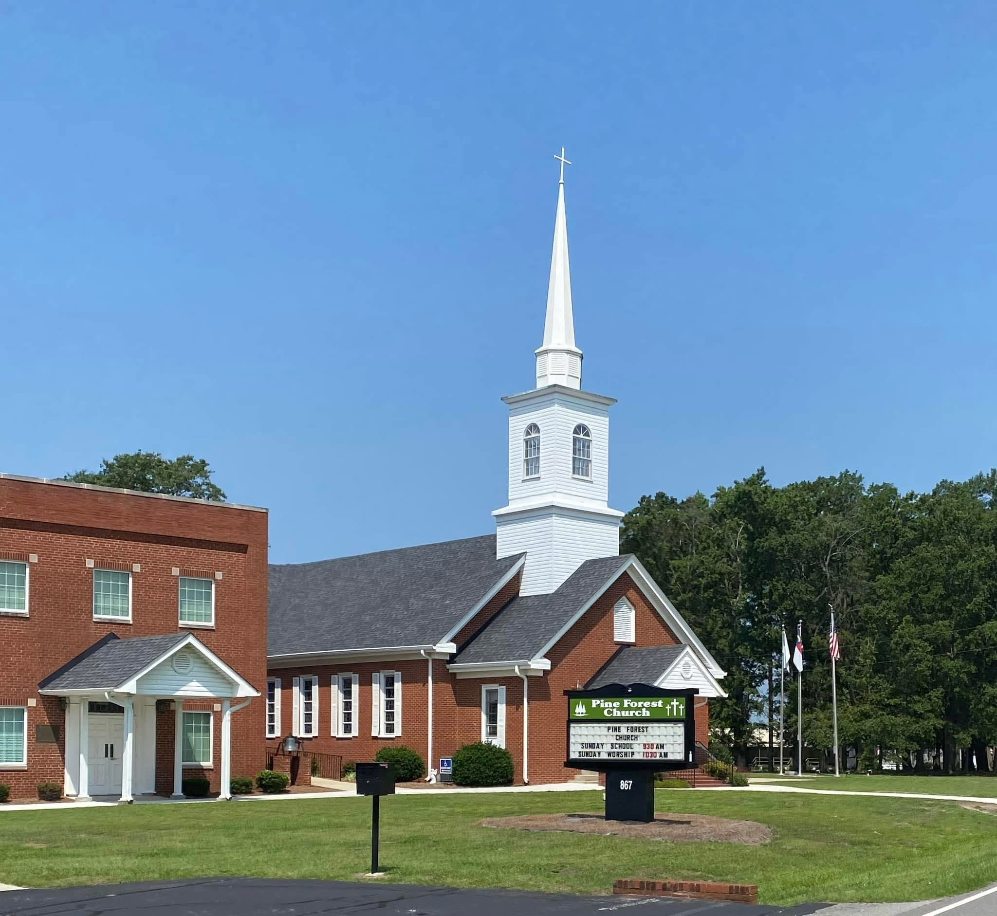 Pine Forest Church features a white steeple and red brick buildings under a clear blue sky. The sign displays service times and church name.