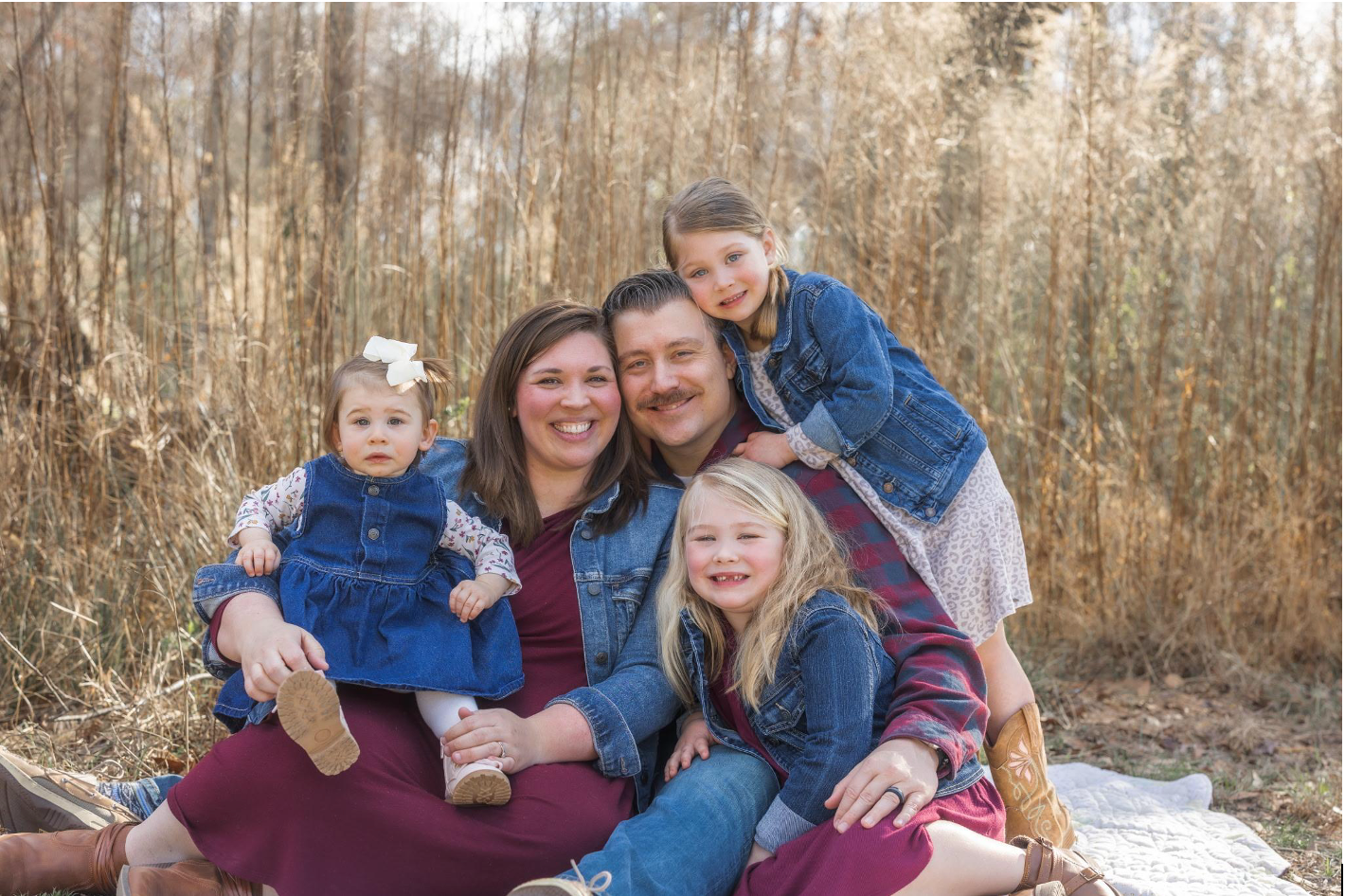 A smiling family of five, wearing coordinated outfits, poses together on a blanket in a wooded area. The image captures a joyful moment and highlights family bonds.