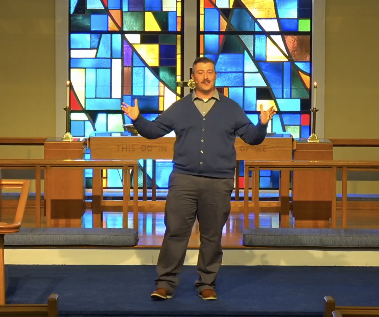 A speaker gestures with open hands in front of a colorful stained glass backdrop in a church setting, engaging the audience.
