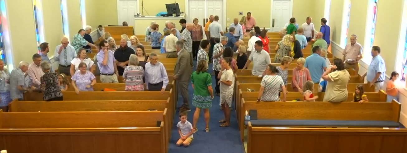 A crowd gathers inside a church during a social event, with individuals mingling and children seated in the pews. Stained glass windows add color.